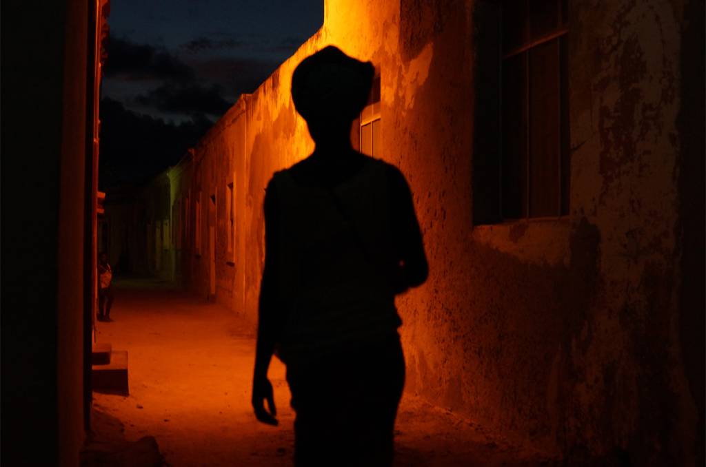 Foto colorida, na foto é possível ver a silhueta de uma pessoa caminhando na rua durante a noite. Logo em frente a ela existe uma luz laranja.
