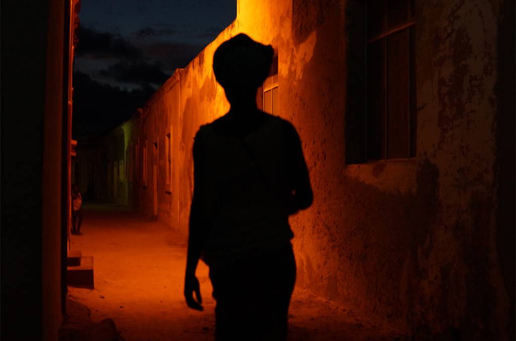 01 Foto colorida, na foto é possível ver a silhueta de uma pessoa caminhando na rua durante a noite. Logo em frente a ela existe uma luz laranja.