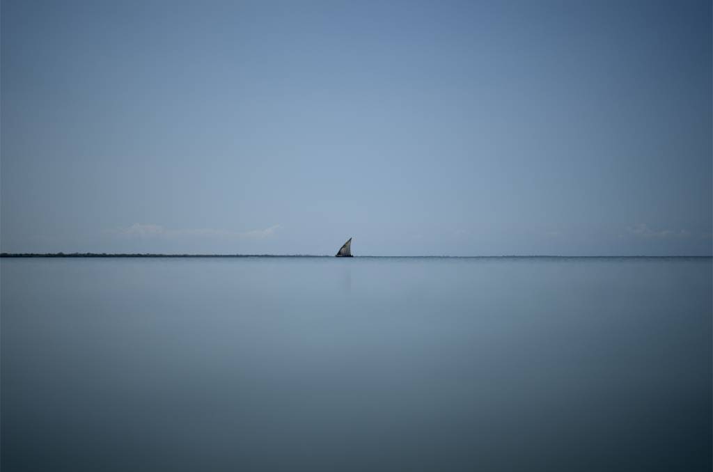 02 Fotografia colorida, na imagem o horizonte, mar e céu. Centralizado entre os dois é possível ver um barco que parece pequeno pela distância.