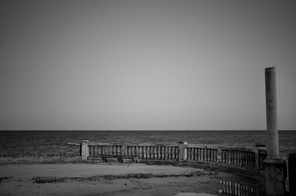 Fotografia preto e branco, na imagem é possível o chão de pedra, uma mureta, o mar e o ceú.
