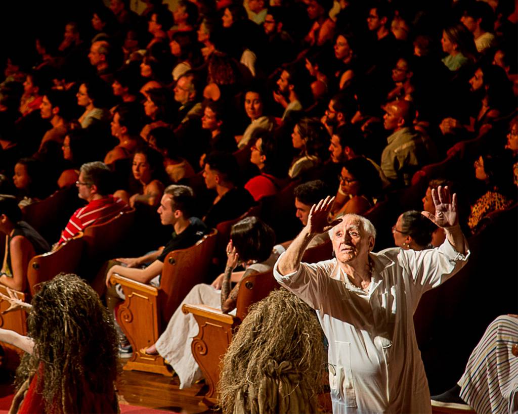 O diretor Zé Celso aparece a direita com as mãos levantadas, atrás dele uma plateia sentada.