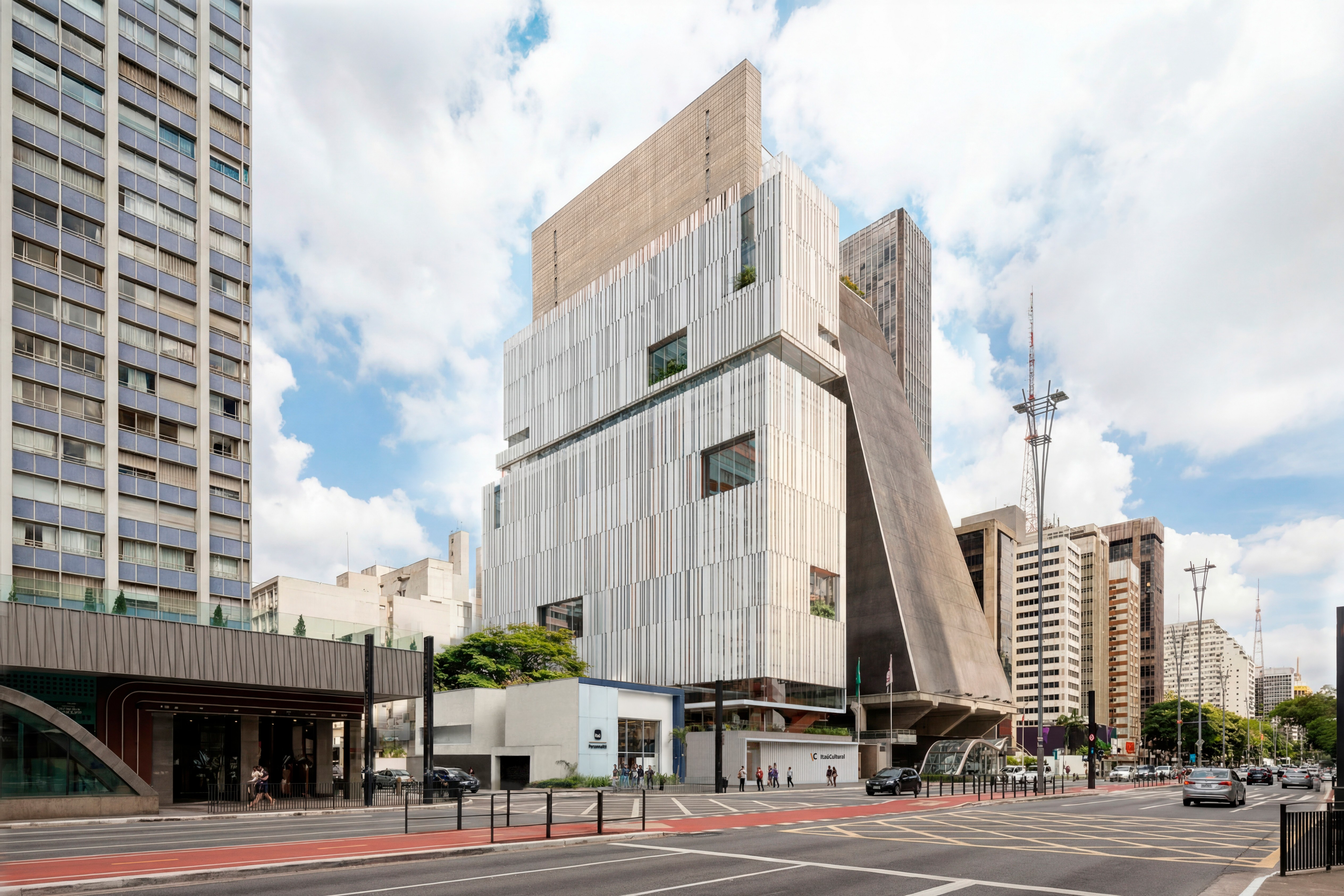 Edifícios modernos e antigos em uma rua movimentada de São Paulo, com céu azul e nuvens. À esquerda, um prédio residencial alto de cor azul e branco. No centro, um edifício branco com fachada verticalmente listrada e janelas quadradas, ao lado de um prédio com fachada inclinada de concreto escuro. Pessoas caminham na calçada e carros circulam na rua com faixa vermelha para ônibus