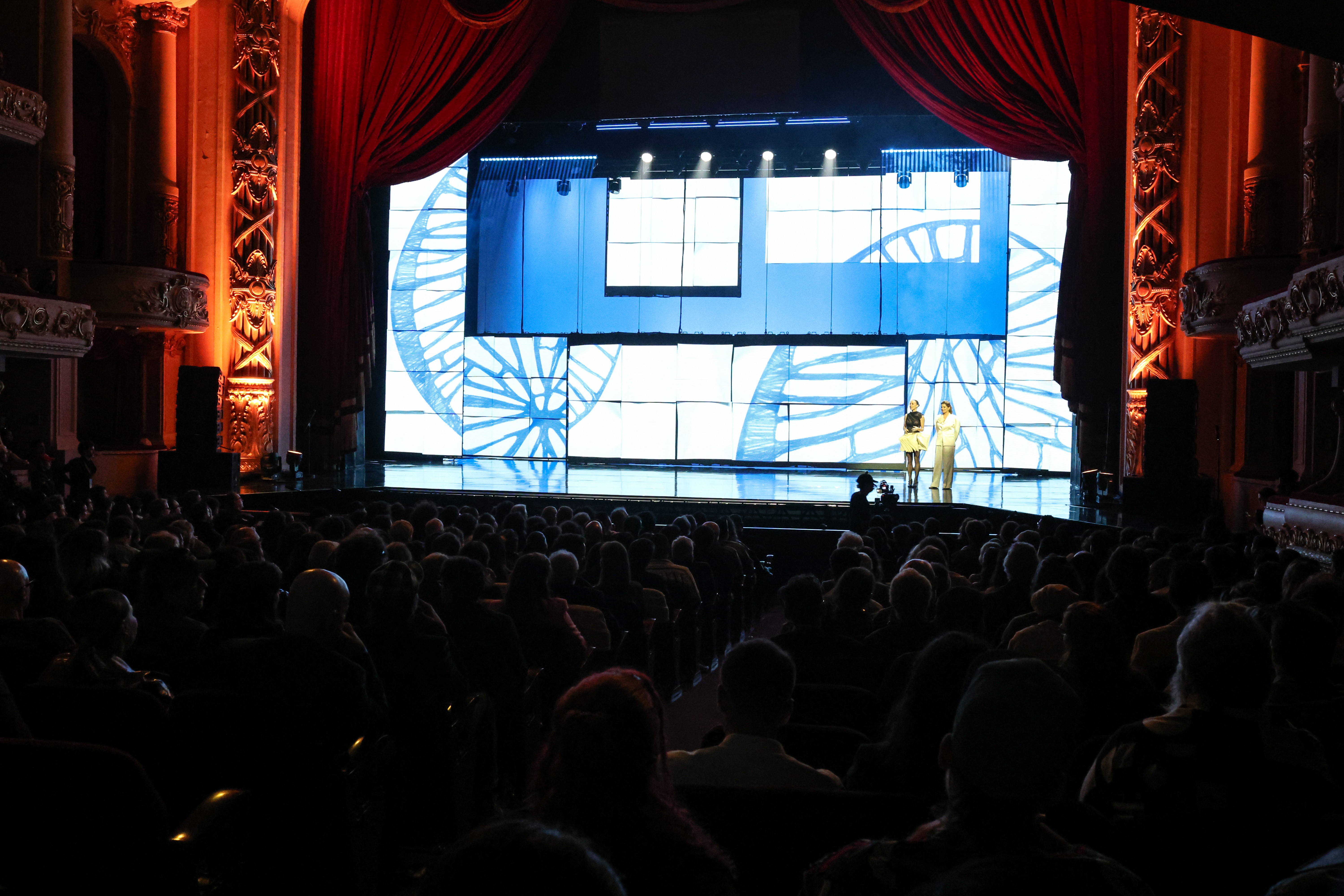 Pessoas assistem a um evento em um teatro com palco iluminado por telas azuis e brancas exibindo desenhos abstratos. Duas mulheres, uma de roupa escura e outra clara, estão no palco. Cortinas vermelhas e detalhes dourados adornam as laterais do palco.