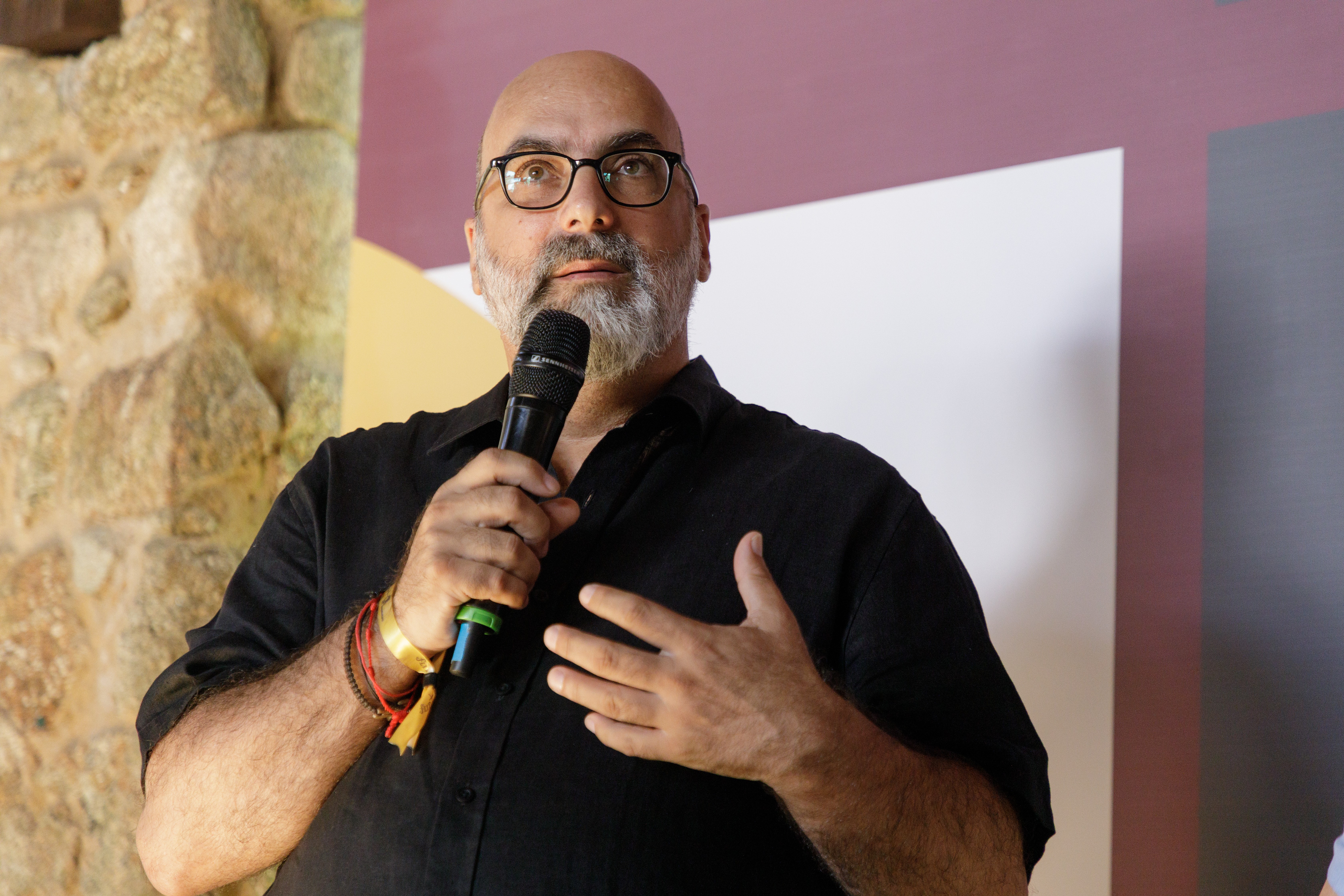Homem calvo de barba grisalha, óculos, camisa preta, segurando um microfone com a mão direita e gesticulando com a esquerda, em um evento com parede de pedra e painéis coloridos