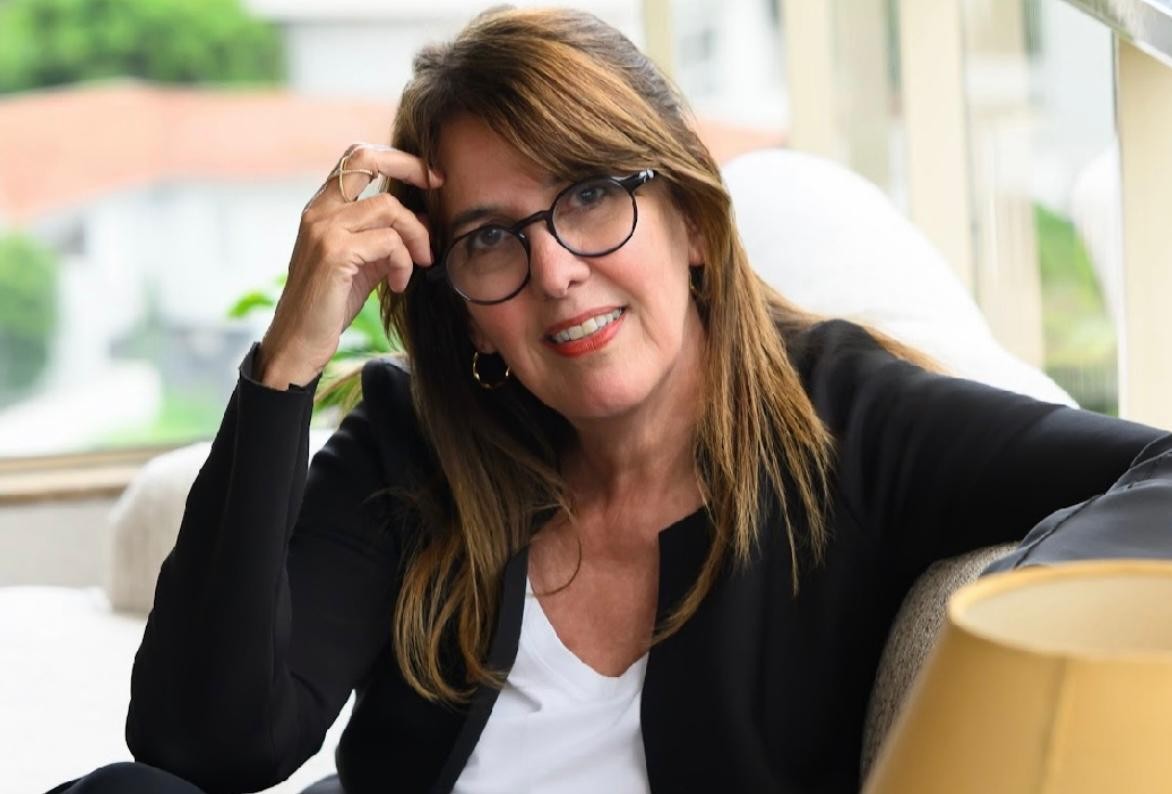 Mulher de meia-idade, cabelos castanhos e óculos, sorri levemente enquanto apoia a mão direita na testa. Veste blazer preto sobre camiseta branca e brincos de argola dourados. Sentada em um sofá claro, com uma luminária amarela no canto inferior direito, e um fundo desfocado de vegetação e telhados