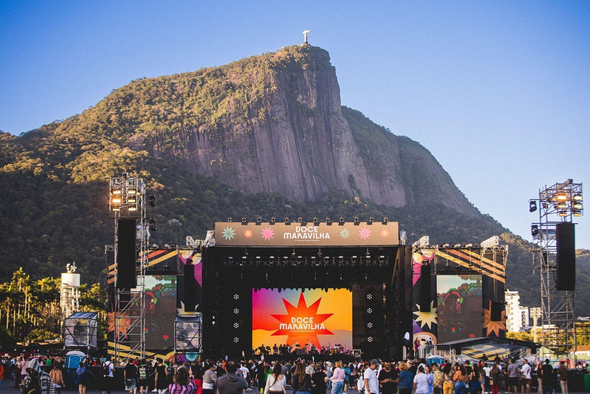 Palco de festival Doce Maravilha com telões coloridos e público, tendo ao fundo o Corcovado com o Cristo Redentor sob céu azul