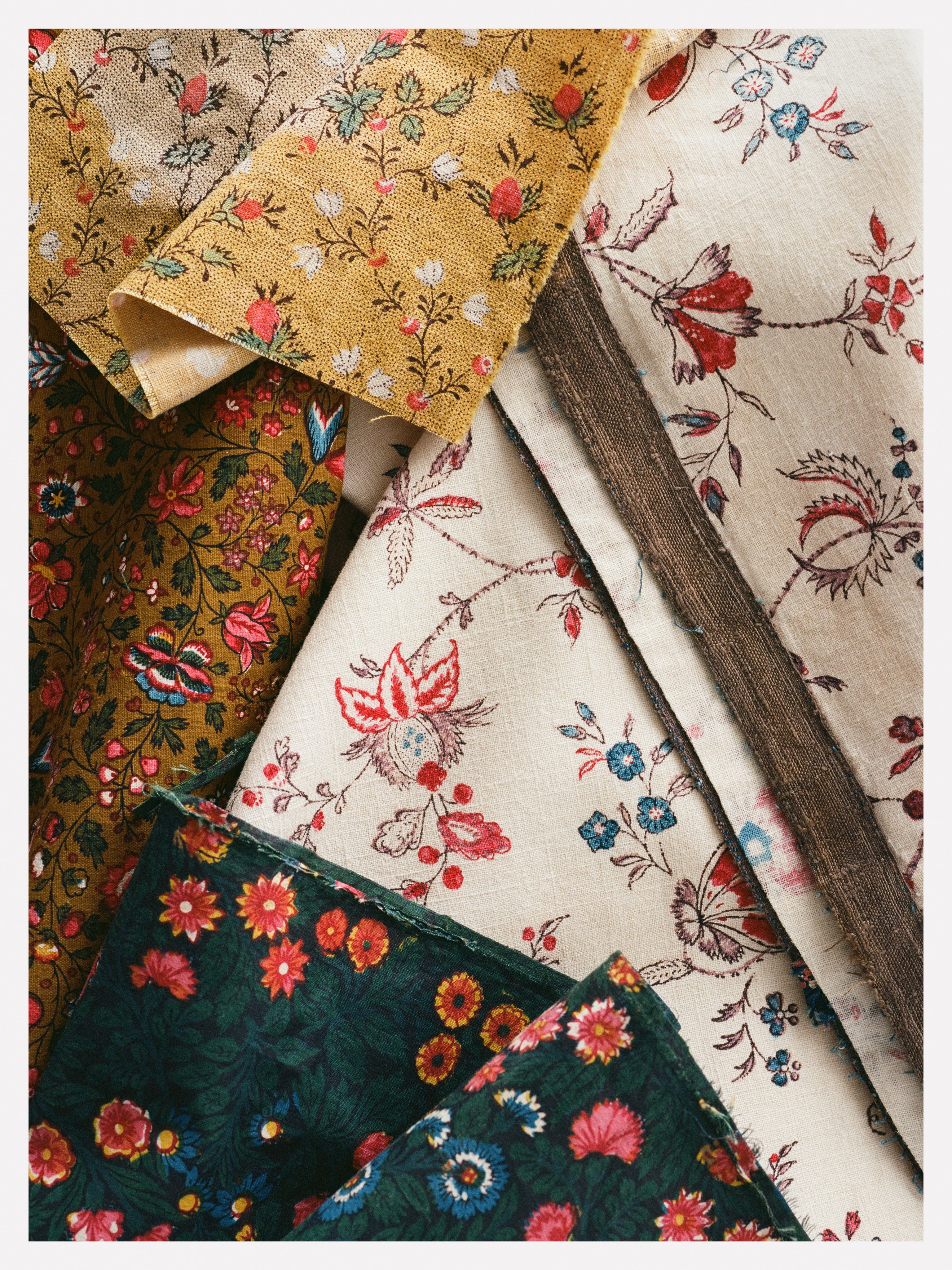 Vários tecidos estampados com motivos florais em tons de mostarda, verde-oliva, creme e verde-escuro, dispostos em camadas sobrepostas