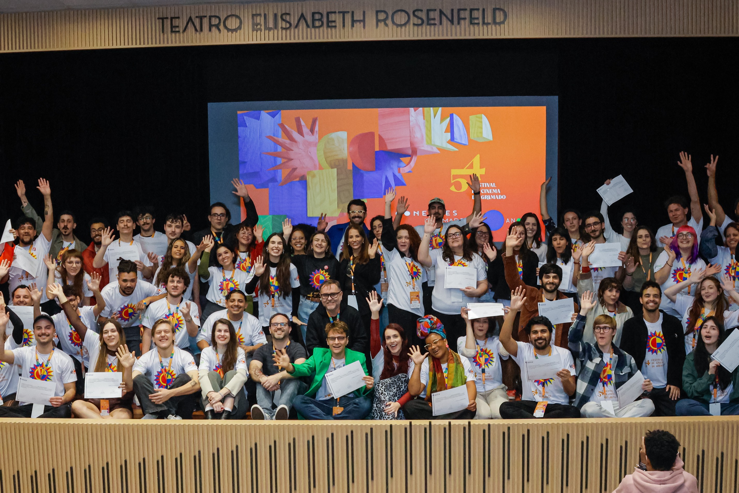 Grande grupo de jovens e adultos, a maioria sorrindo e acenando, alguns segurando certificados, em frente a um palco com a inscrição TEATRO ELISABETH ROSENFELD e um telão exibindo a logo do 51º FESTIVAL DE CINEMA DE GRAMADO