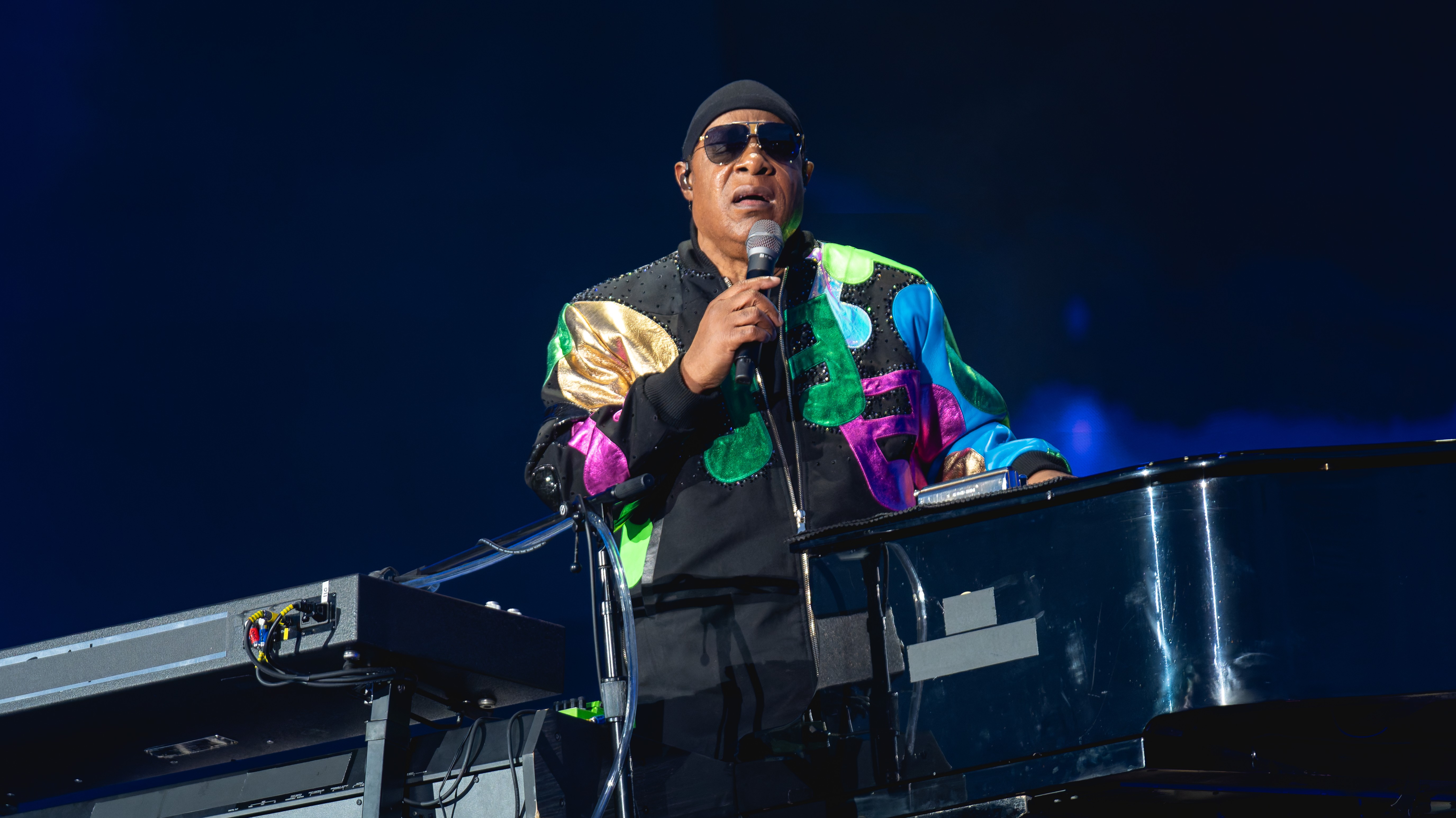 Stevie Wonder, usando óculos escuros, gorro preto e jaqueta colorida com detalhes brilhantes, canta em um microfone enquanto toca piano em um palco escuro