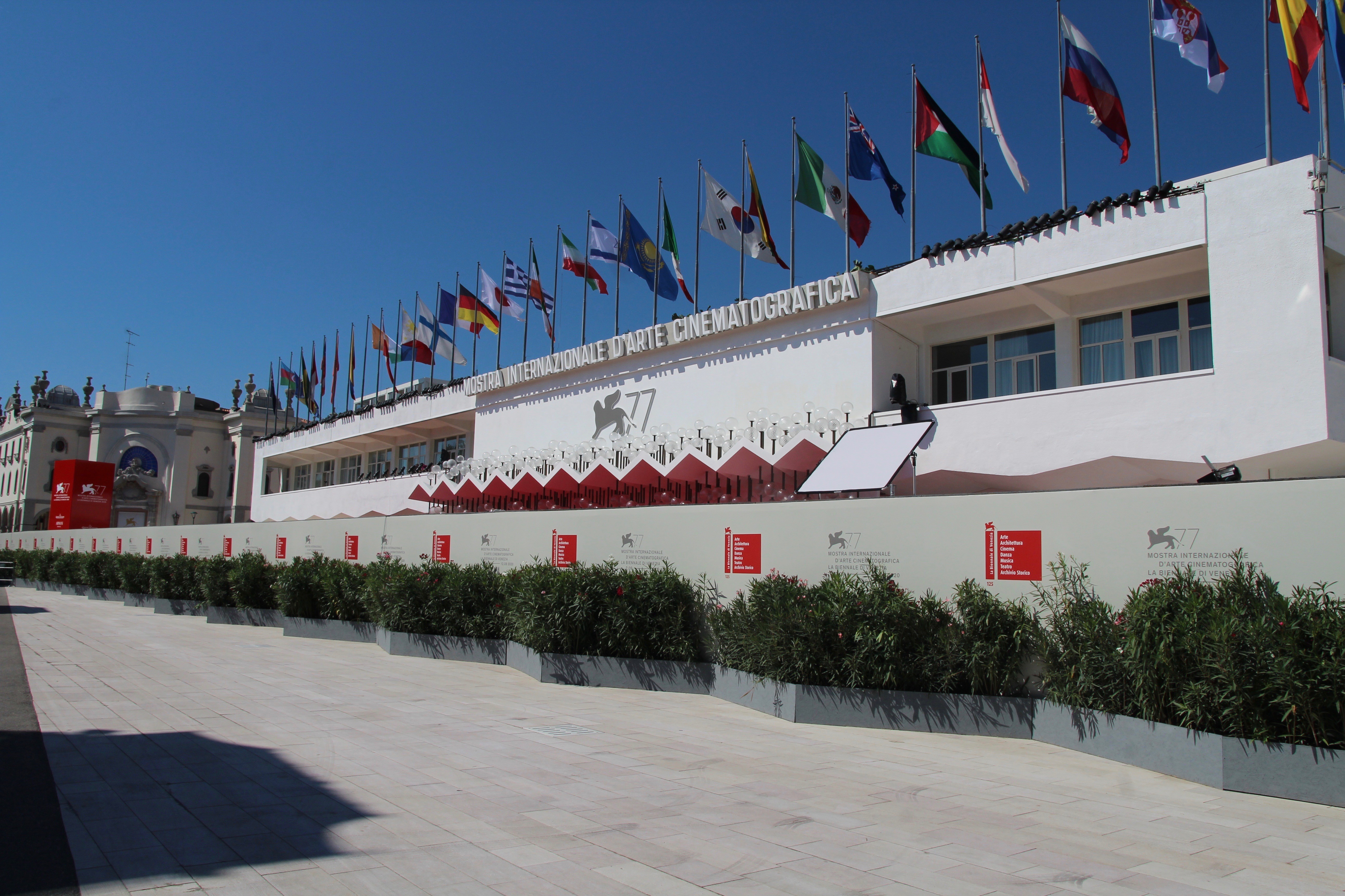 Fachada branca do Palazzo del Cinema em Veneza, com a inscrição MOSTRA INTERNAZIONALE DARTE CINEMATOGRAFICA e o logo da 77ª edição. Várias bandeiras de países tremulam no alto, sob um céu azul claro. Em primeiro plano, canteiros com arbustos verdes e um calçadão de pedra. À esquerda, parte de um edifício clássico.