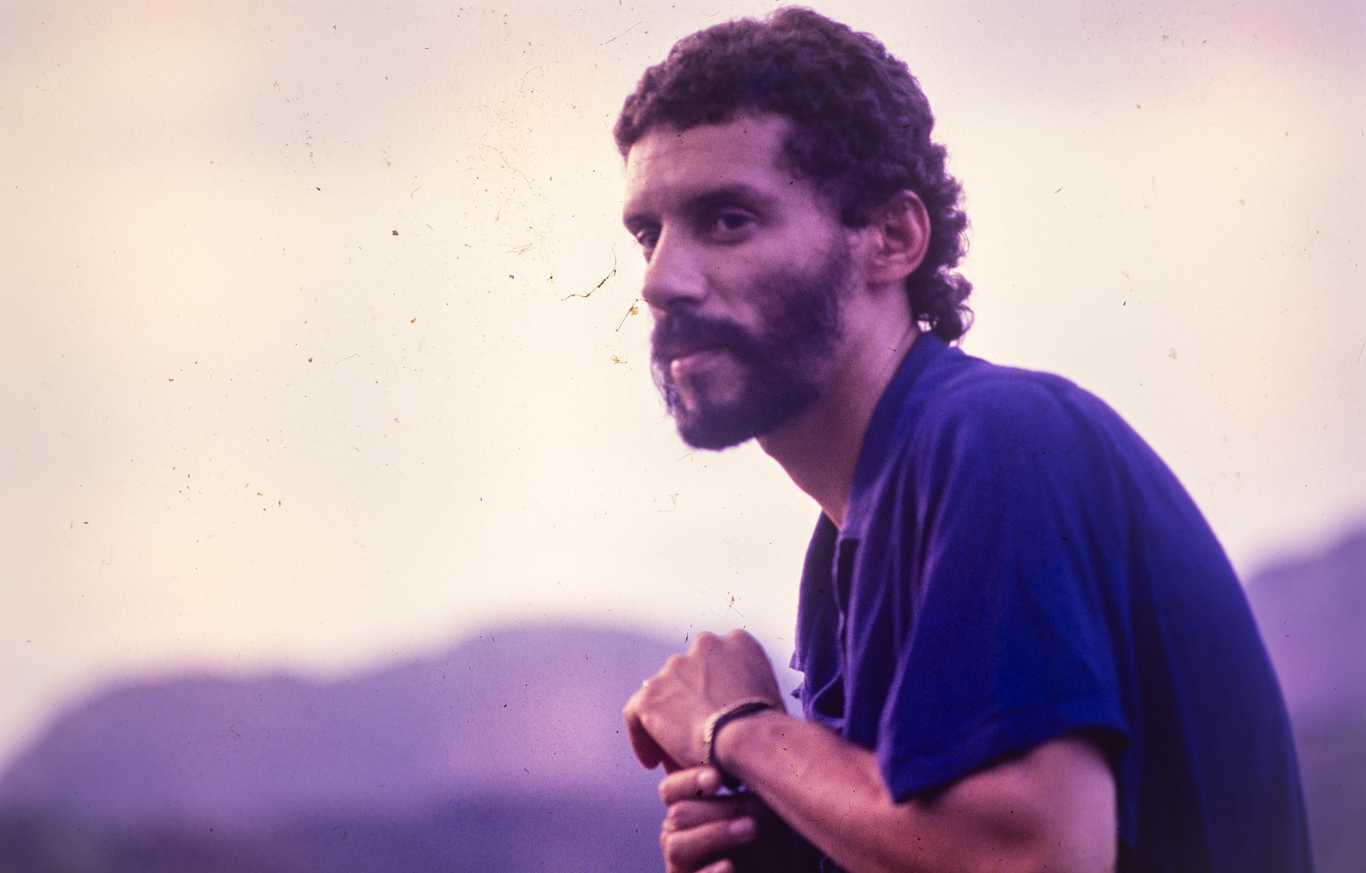 Homem de pele clara, cabelo e barba crespos escuros, vestindo camisa polo azul, olhando para a direita com expressão pensativa. Ao fundo, montanhas em tons de roxo e céu claro.