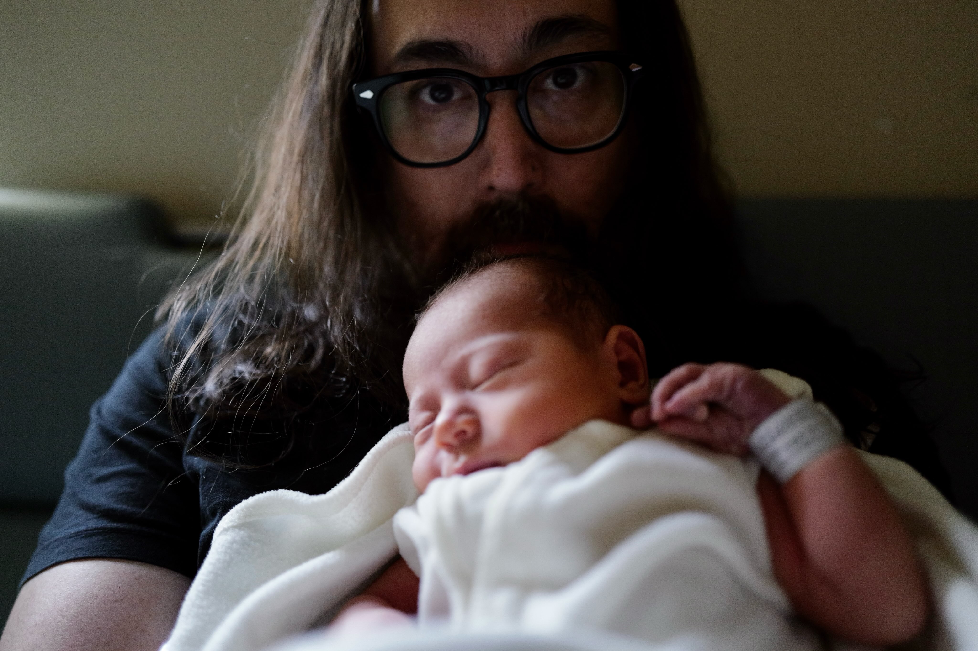 Um homem de cabelo e barba longos, usando óculos escuros e camiseta preta, segura um bebê recém-nascido adormecido, enrolado em um cobertor branco. O bebê tem uma pulseira de identificação no pulso esquerdo. O homem olha diretamente para a câmera com uma expressão séria
