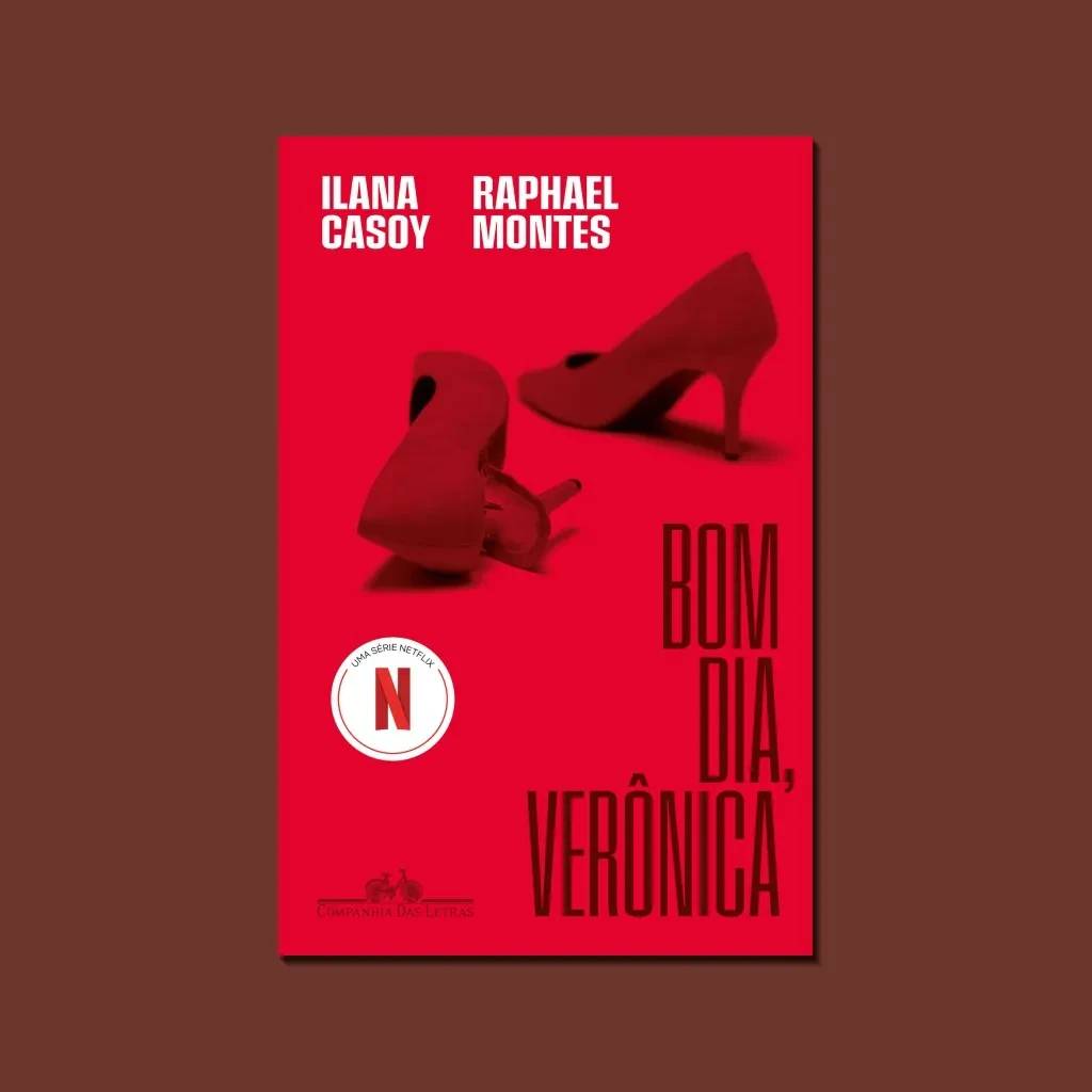 Capa do livro Bom Dia, Ver&ocirc;nica com fundo vermelho vibrante. Dois sapatos de salto alto vermelhos, um deitado e outro em p&eacute;, ocupam o centro. No topo, os nomes dos autores Ilana Casoy e Raphael Montes. Abaixo, o t&iacute;tulo em letras grandes. Um selo circular branco com o logo da Netflix e a frase Uma S&eacute;rie Netflix aparece no canto inferior esquerdo. Na base, o logo da Companhia das Letras