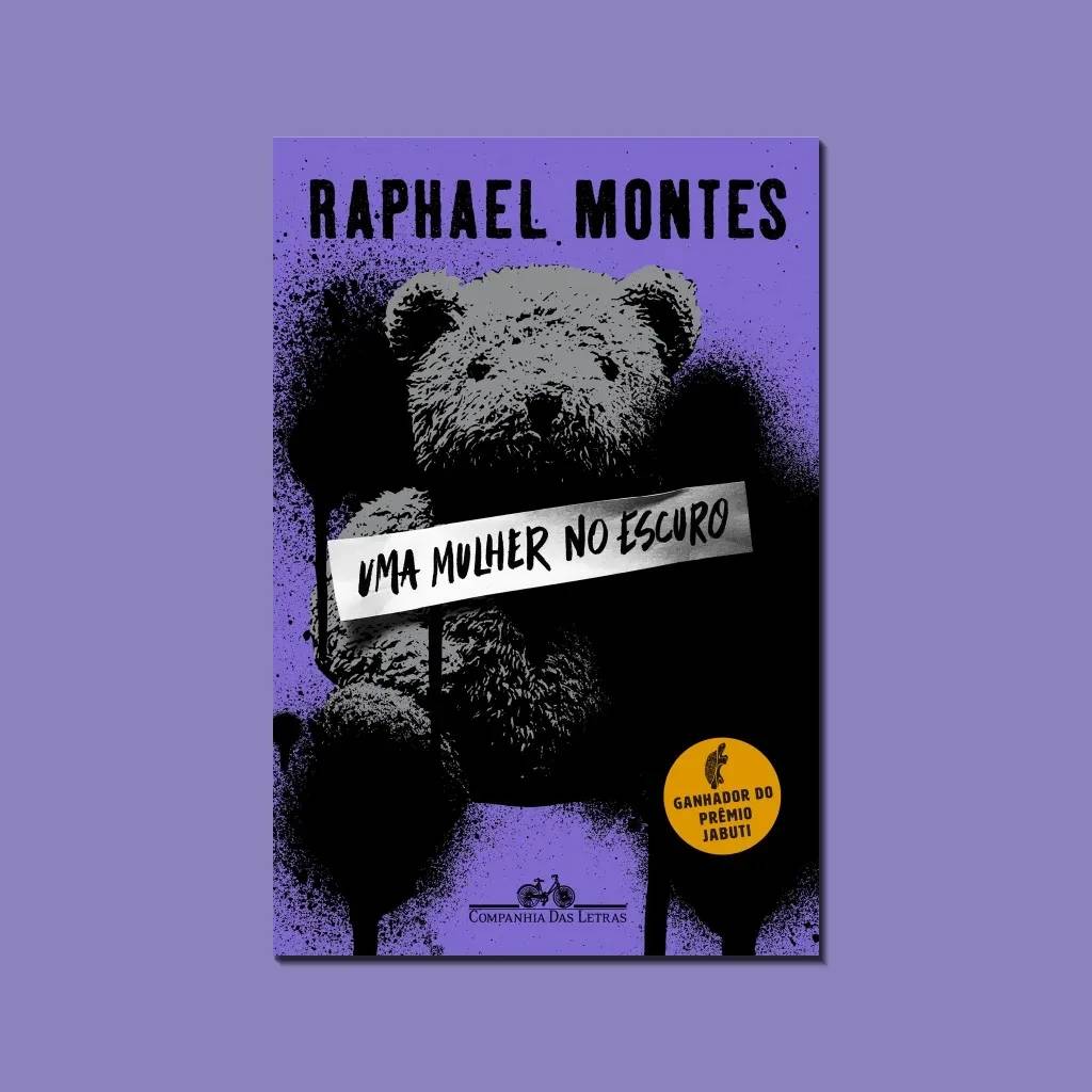 Capa do livro Uma Mulher no Escuro de Raphael Montes. Fundo roxo com um urso de pel&uacute;cia cinza e preto, como um grafite. Uma faixa branca com o t&iacute;tulo em preto atravessa o peito do urso. No canto inferior direito, um selo amarelo indica Ganhador do Pr&ecirc;mio Jabuti. A editora Companhia das Letras aparece na parte inferior