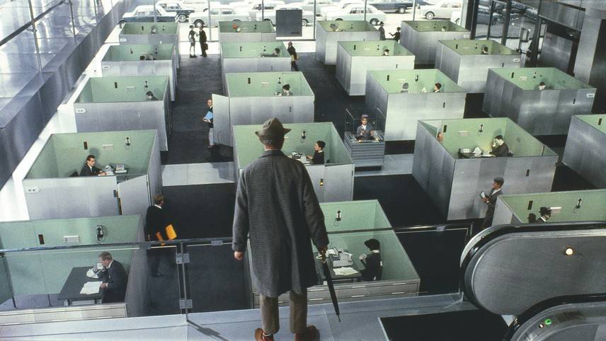 Homem de chap&eacute;u e sobretudo cinza, de costas, observa um escrit&oacute;rio futurista com cub&iacute;culos individuais, onde pessoas trabalham em mesas com telefones. Uma escada rolante aparece &agrave; direita e carros estacionados ao fundo, vis&iacute;veis atrav&eacute;s de uma grande janela