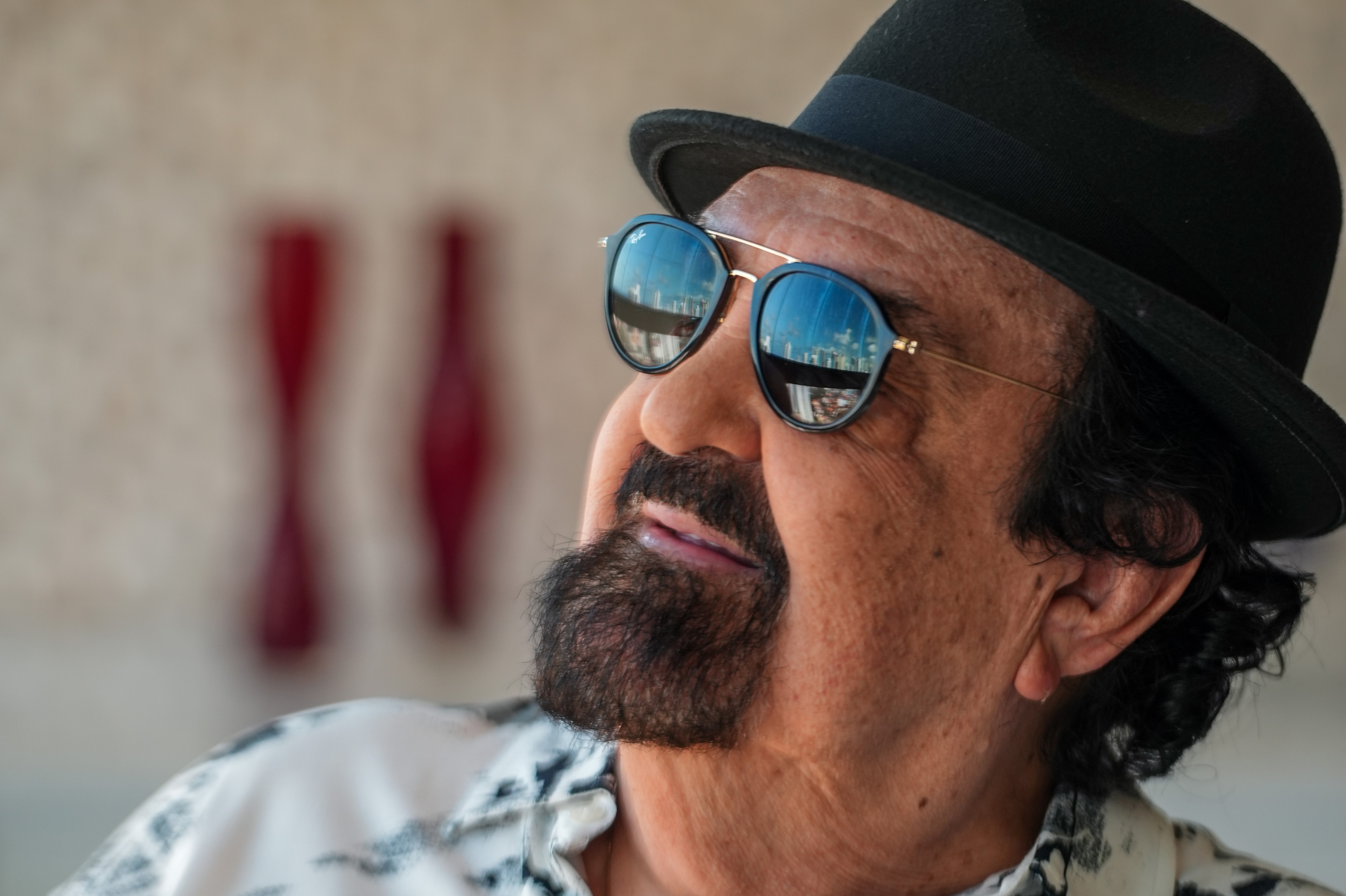 Homem de pele morena, barba preta e chapéu fedora escuro, usando óculos de sol que refletem edifícios, olhando para cima com expressão pensativa. Veste camisa estampada em tons claros.