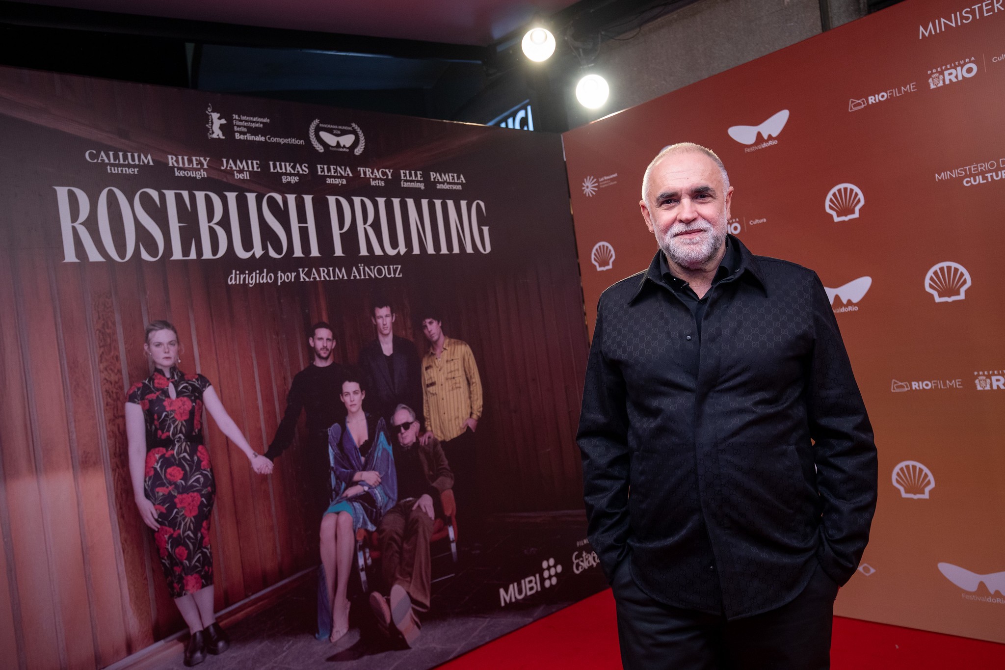 Homem de barba grisalha e camisa preta texturizada posa sorridente em frente a um painel vermelho com logos e um pôster do filme Rosebush Pruning