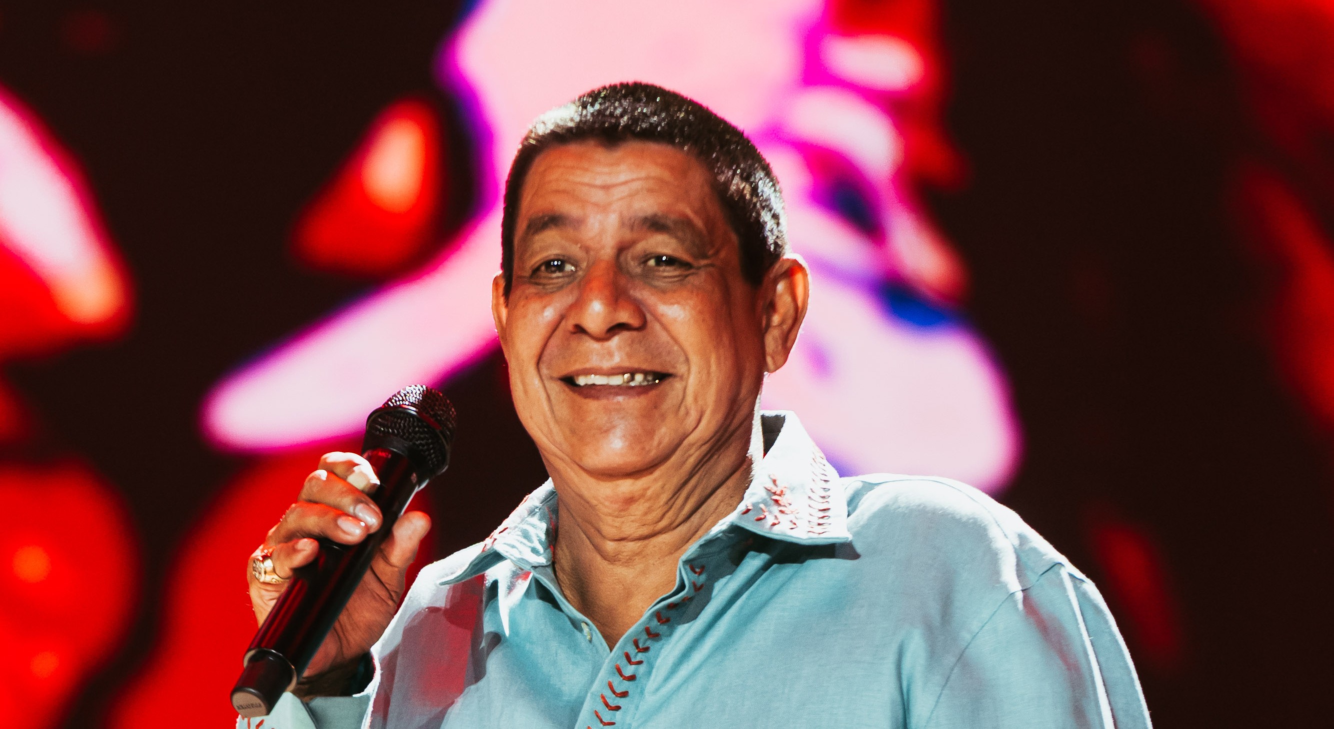 Um homem de pele morena, cabelo escuro e curto, sorri amplamente enquanto segura um microfone preto na mão direita. Ele veste uma camisa azul-clara com detalhes bordados em vermelho no colarinho e na manga. O fundo está desfocado com luzes vermelhas e roxas
