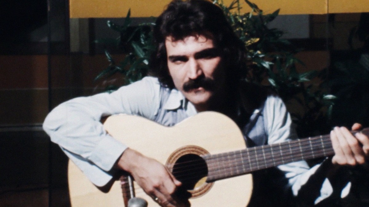 Homem de cabelos e bigode escuros, vestindo camisa clara, tocando violão com as mãos sobre as cordas, em ambiente com plantas e fundo escuro