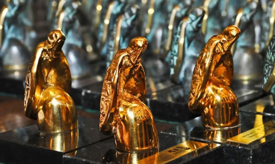 Três estatuetas douradas em primeiro plano, representando figuras sentadas com capuzes, sobre bases pretas. Ao fundo, outras estatuetas similares em tons esverdeados, desfocadas.
