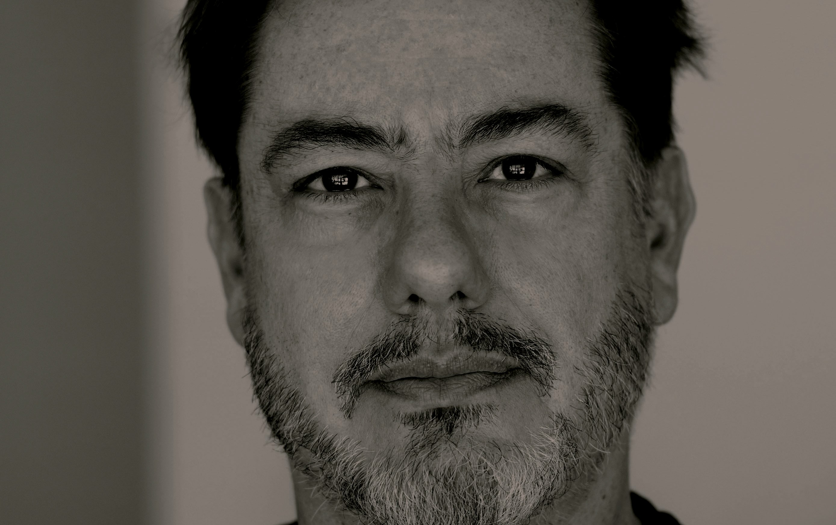 Rosto de um homem de meia-idade, em preto e branco, com barba e cabelo escuros, olhando diretamente para a câmera com expressão séria