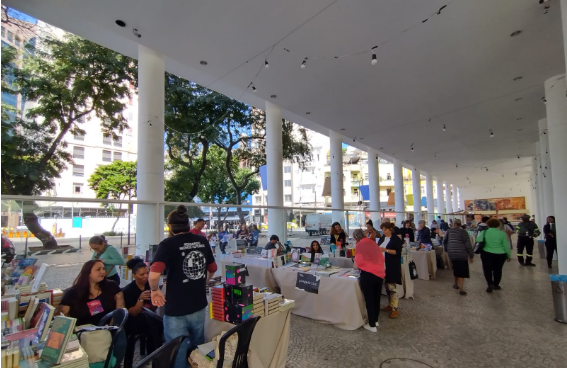 Pessoas em uma feira de livros ao ar livre, com mesas cobertas por toalhas claras exibindo publicações. Ao fundo, prédios e árvores verdes sob um céu azul.